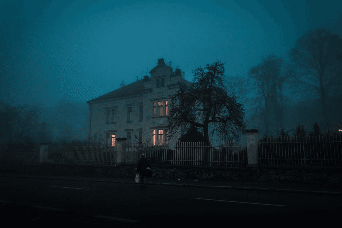 A fog-covered old house with warm lights glowing through its windows at dusk. A moody cinematic autumn night scene.